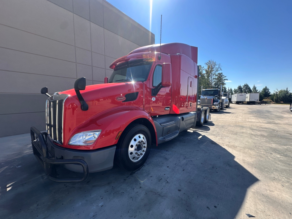 Peterbilt 579 conventional sleeper truck in red, front three-quarters view, with chrome grille, black bumper, dual rear wheels, daytime sunlight.
