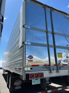 Rear view of a 2018 Utility 3000R W trailer showing steel roll-up doors, taillights, bumper, and Utility 3000R badge on the lower left; reflective surface and light blue sky.