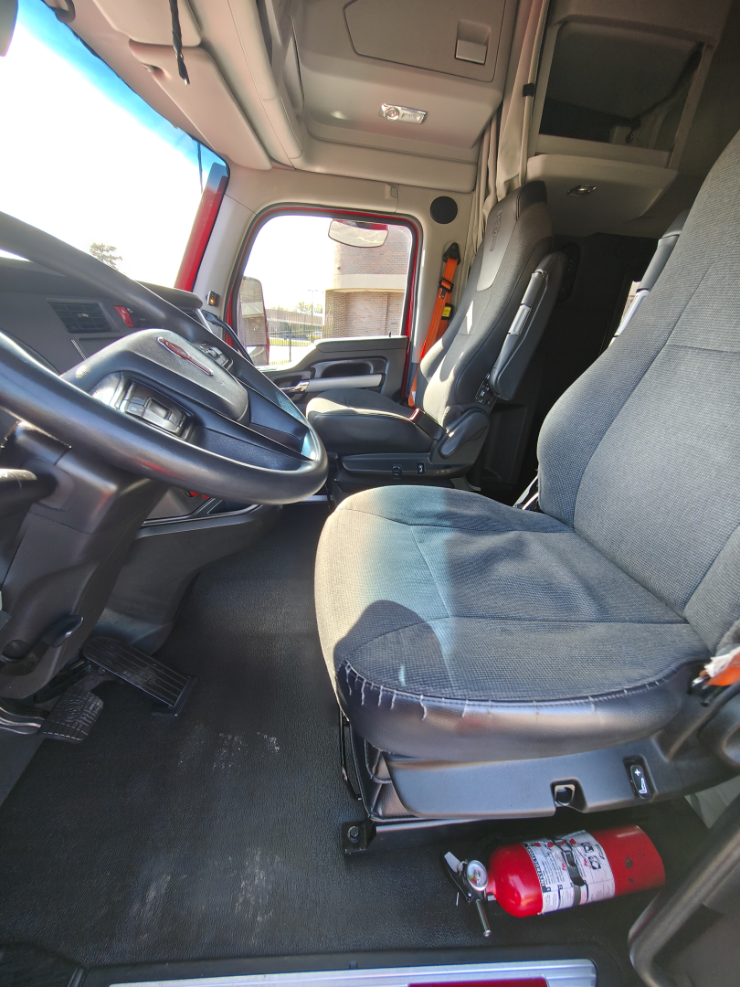 Kenworth T680 truck cab interior showing driver's seat, steering wheel, and dash with a red fire extinguisher on the floor near the seat.