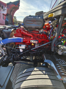 Kenworth T680 engine bay with bright red engine labeled 6x15 and blue silicone hose; visible battery box and wiring, hood open