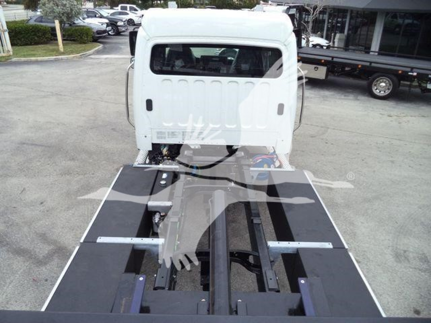Truck cab exposed from rear showing a flatbed/frame of a 2027 Freightliner M2 106 rollback chassis, white cab with black platform, engine bay hidden, dealership lot in background.