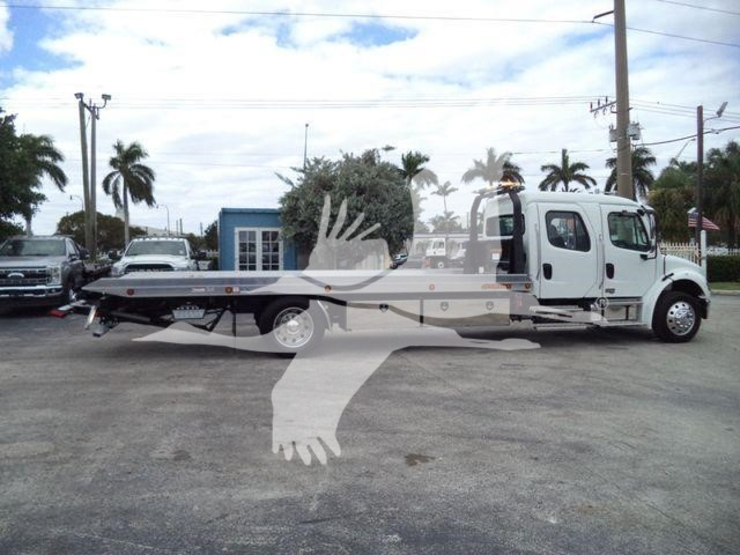 Freightliner M2 106 rollback truck side view showing flatbed, cab, and up-front wheel; visible dealership lot in background.