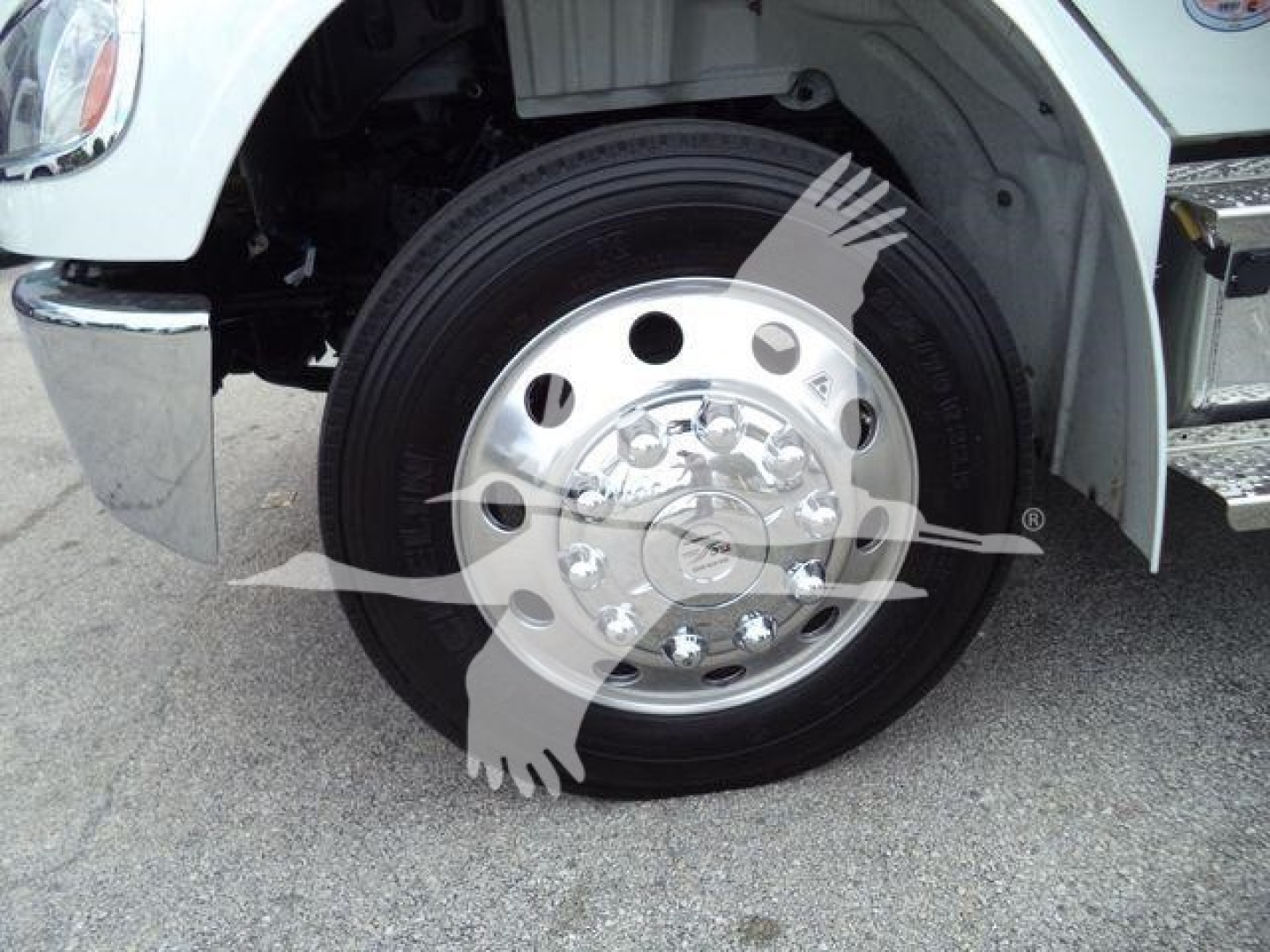 Front wheel and fender of a Freightliner M2 106 truck showing a chrome hubcap on a black tire, with part of the bumper and wheel well visible.