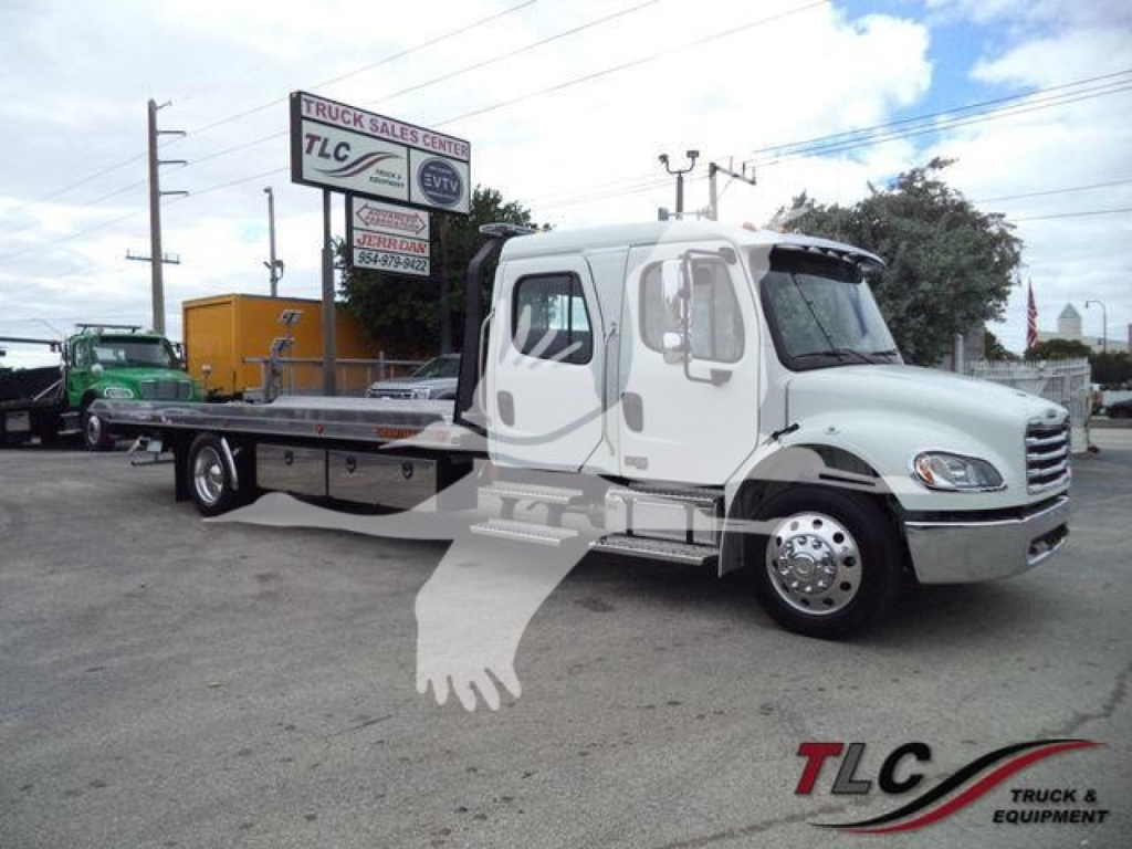 Freightliner M2 106 rollback truck in a lot; white cab with gray flatbed; TLC Truck Sales Center sign in background; JU6165 stock number; rolling equipment visible.