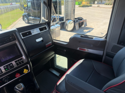 Kenworth W900L truck cab interior view showing driver seat, dashboard with controls, and large side mirror; window reflects other trucks in lot.