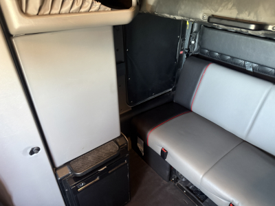 Kenworth W900L cab interior showing sleeper area and seating with light gray upholstery and red piping, compact fridge visible near left wall, black cabinet and door in background.