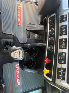 Kenworth W900L dash area showing control panel and Kelley Kenworth branding on floor mats, gear shifter visible, various knobs and switches.