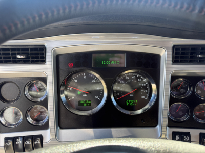 Kenworth W900L truck cockpit dashboard view showing speedometer and tachometer gauges, center digital clock, and surrounding gauges.