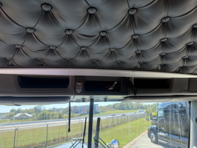Kenworth W900L truck interior ceiling view with tufted black upholstery and dashboard vents, windshield showing highway and truck parking area.