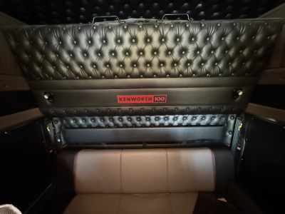 Kenworth W900L sleeper cab interior with quilted black upholstery and a Kenworth 100 emblem on the panel above a beige seat cushion.