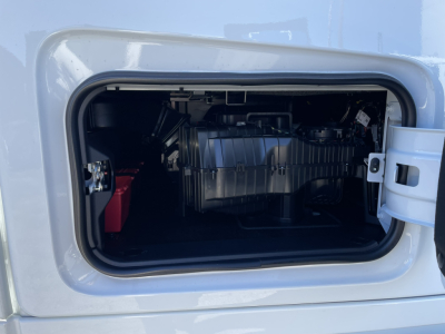 Truck engine bay access door open showing battery on left, engine components, and a metallic intake housing across the middle.