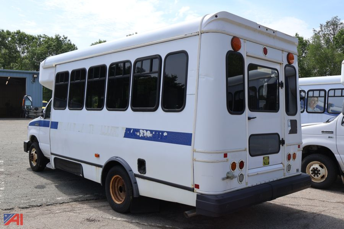 Ford E-350 shuttle bus rear side view showing wheelchair bus configuration, white with blue stripe, multiple windows, rear bumper and spare tire mount.