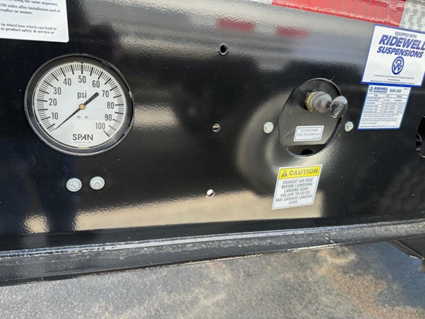 Pressure gauge labeled 'SPAN' shows 0-100 psi scale; various stickers and warning labels on a black panel; circular connector or valve with a label; blue Ridewell suspensions sticker; yellow caution label; part of a dump trailer dashboard.