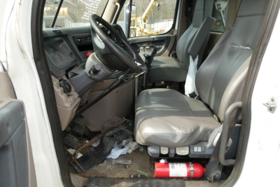 Cab interior of a Freightliner Cascadia 125 conventional daycab showing driver seat, passenger seat, steering wheel, dashboard, and clutter on floor with fire extinguisher under seat.