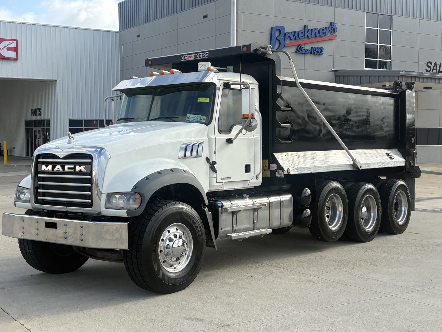 Mack GR64F dump truck in front of dealership, white cab with black dump body, three-axle configuration, chrome grille, visible Mack logo, dealership building in background.