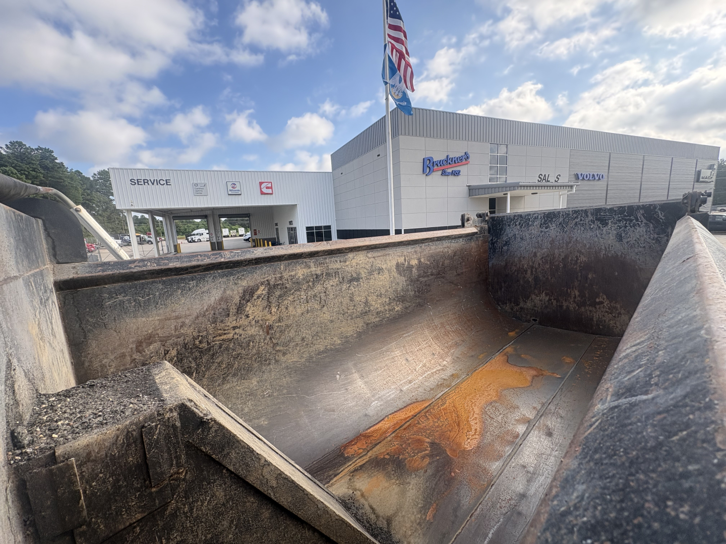 Dump truck bed interior with rust stains and blackened metal; background shows a Mack dealership building with SERVICE sign and American flag.