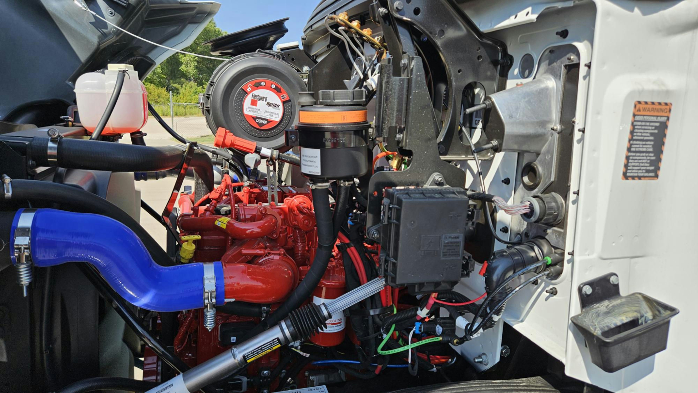 Mack MD6 engine bay showing red engine, blue coolant hose, black air intake, wiring harnesses and white body panel with orange warning label.