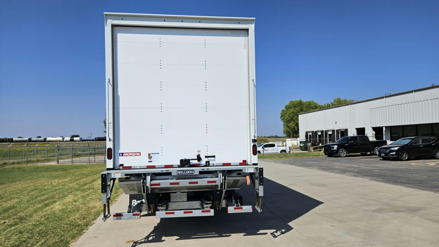Rear view of a 2025 Mack MD6 box truck showing the roll-up cargo door, bumper, taillights, and reflective tape.