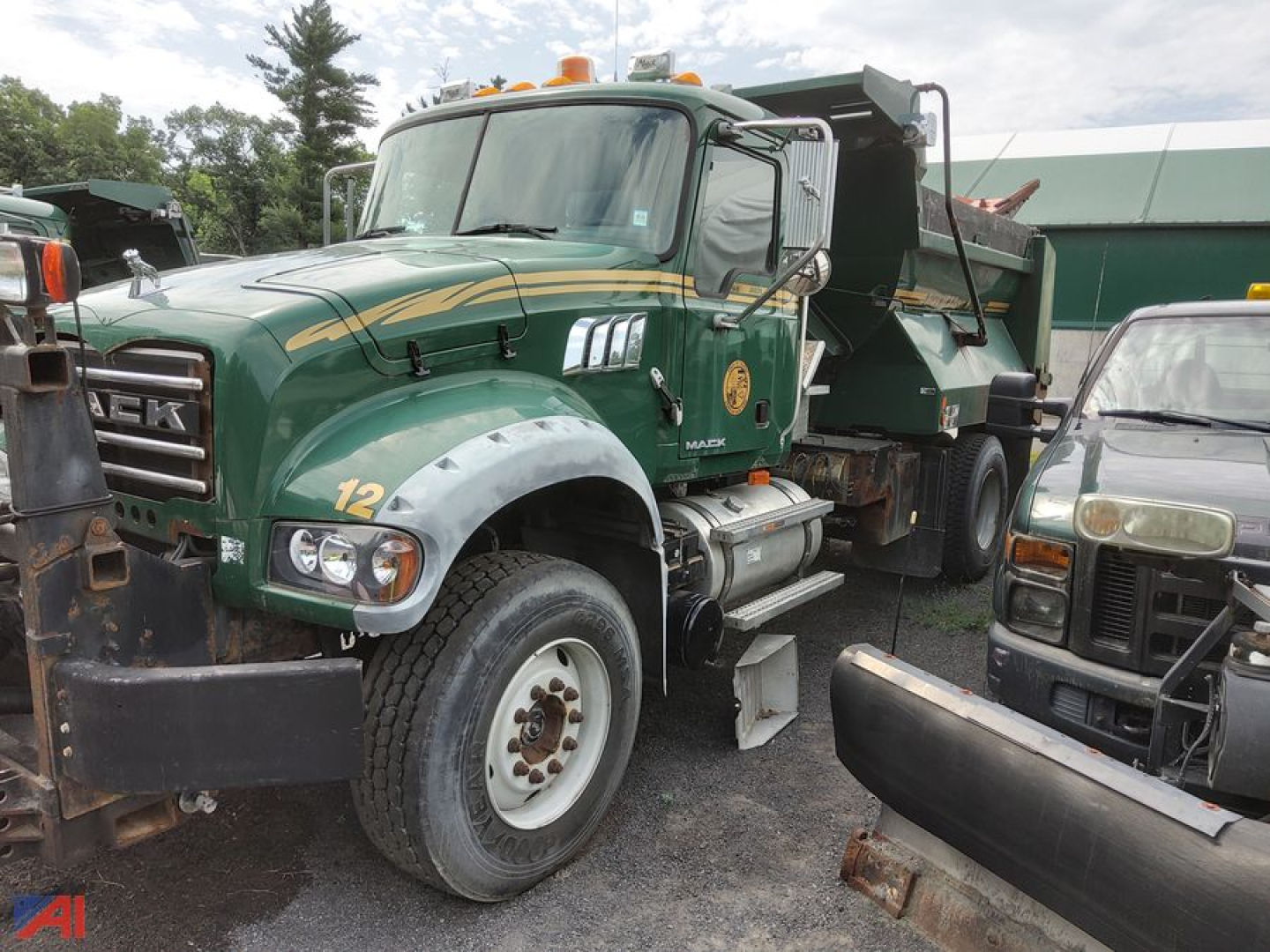 Mack Granite dump truck with plow attachment and green and gold livery, front-left view showing large tires, reflective mirrors, and dump bed