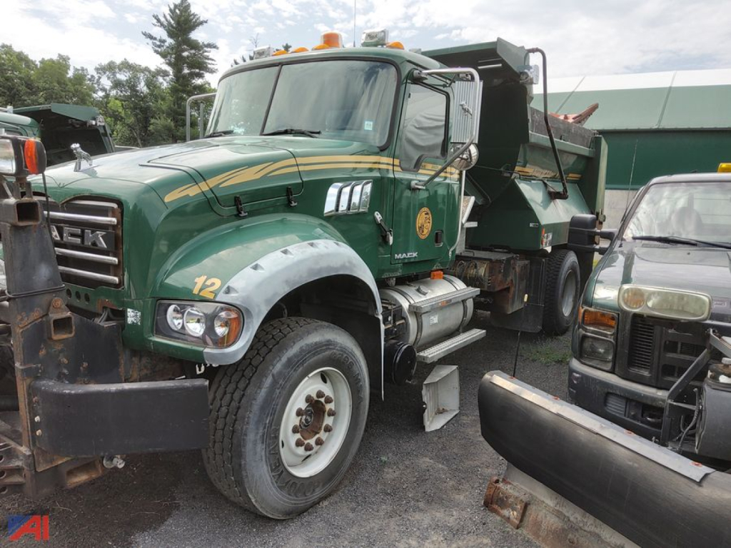 Mack Granite dump truck with plow attachment and green and gold livery, front-left view showing large tires, reflective mirrors, and dump bed