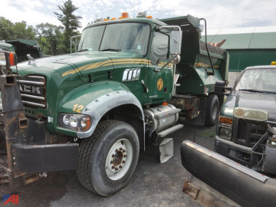 Mack Granite dump truck with plow attachment and green and gold livery, front-left view showing large tires, reflective mirrors, and dump bed