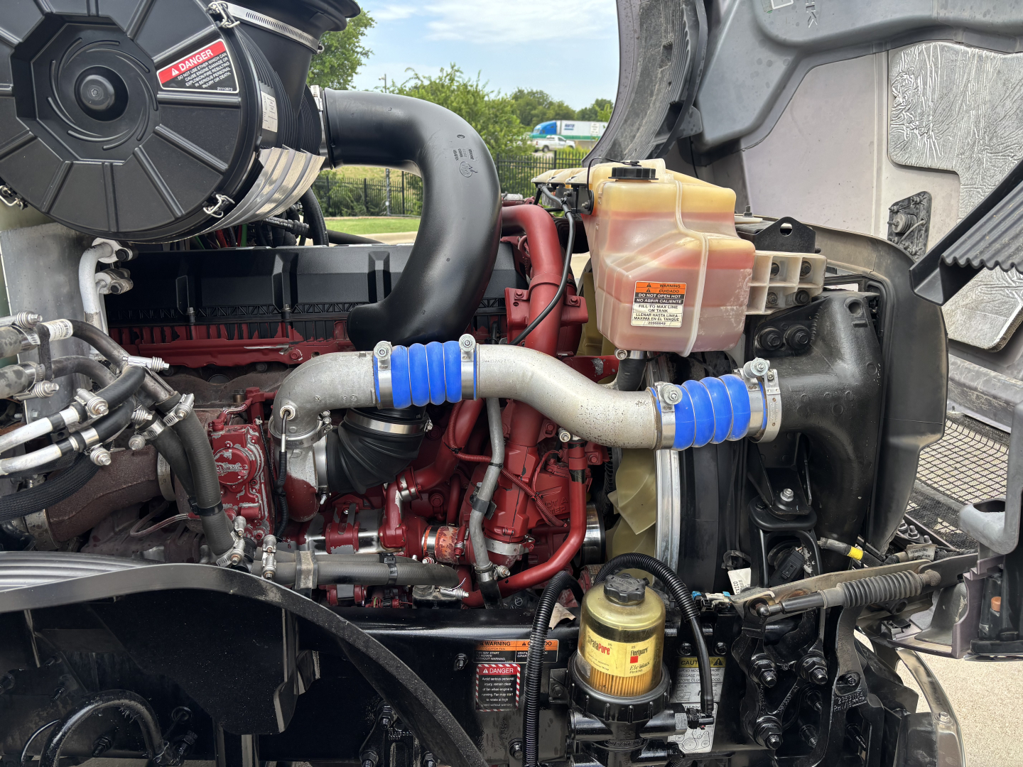 Engine bay of a 2023 Mack AN64T70 truck showing coolant reservoir, hoses, turbo piping, fuel filter housing, and engine block with red paint.