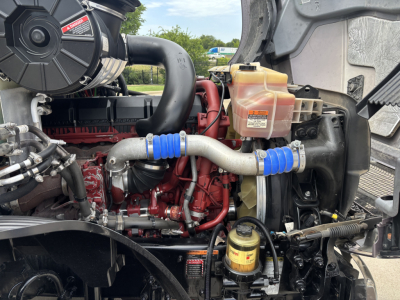 Engine bay of a 2023 Mack AN64T70 truck showing coolant reservoir, hoses, turbo piping, fuel filter housing, and engine block with red paint.
