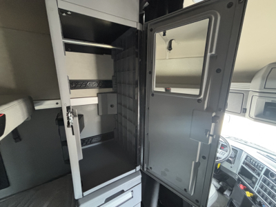 Truck interior showing cab area with tall metal cabinet and open refrigerator/freezer unit inside sleeper berth.