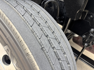 Tire tread close-up on Mack AN64T70 truck; visible text on tire sidewall includes punctured tread pattern and part of wheel hub and suspension components.