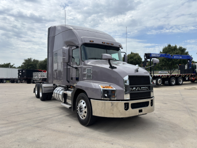 Mack AN64T70 conventional sleeper truck in a parking lot, gray cab with chrome front bumper, Mack logo on grille, side fuel tanks, dual rear axles.