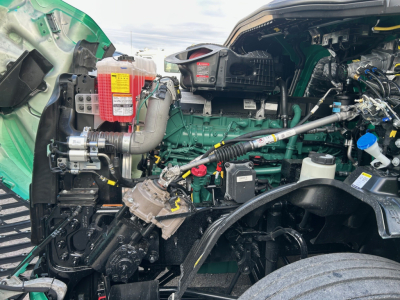 Volvo VNL truck engine bay showing green engine block with red coolant reservoir, black hoses, and yellow warning labels; various components visible in front of cab.