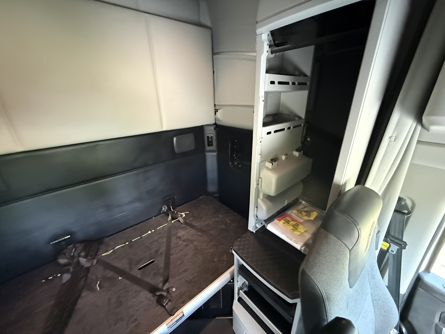 Kenworth T680 truck cab interior view showing storage compartments with shelves and a folded passenger seat; vertical cabinet with small items; light interior panels.