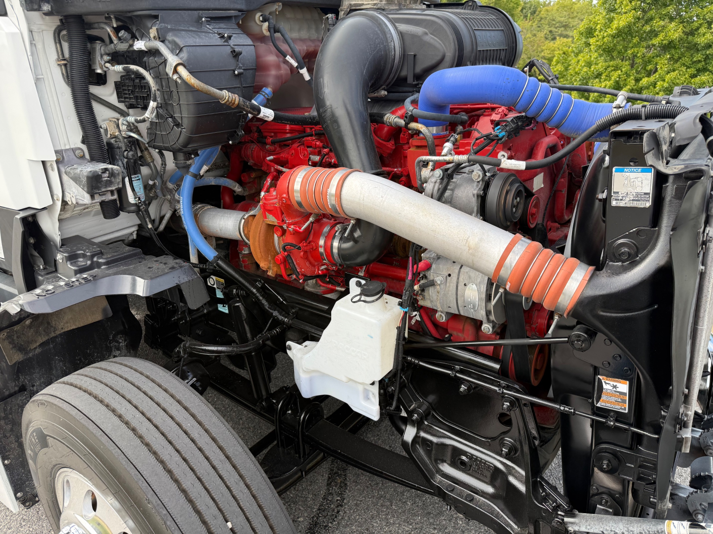 Kenworth T680 engine bay showing red engine block, blue coolant lines, orange intake hose, black chassis, and white coolant reservoir.