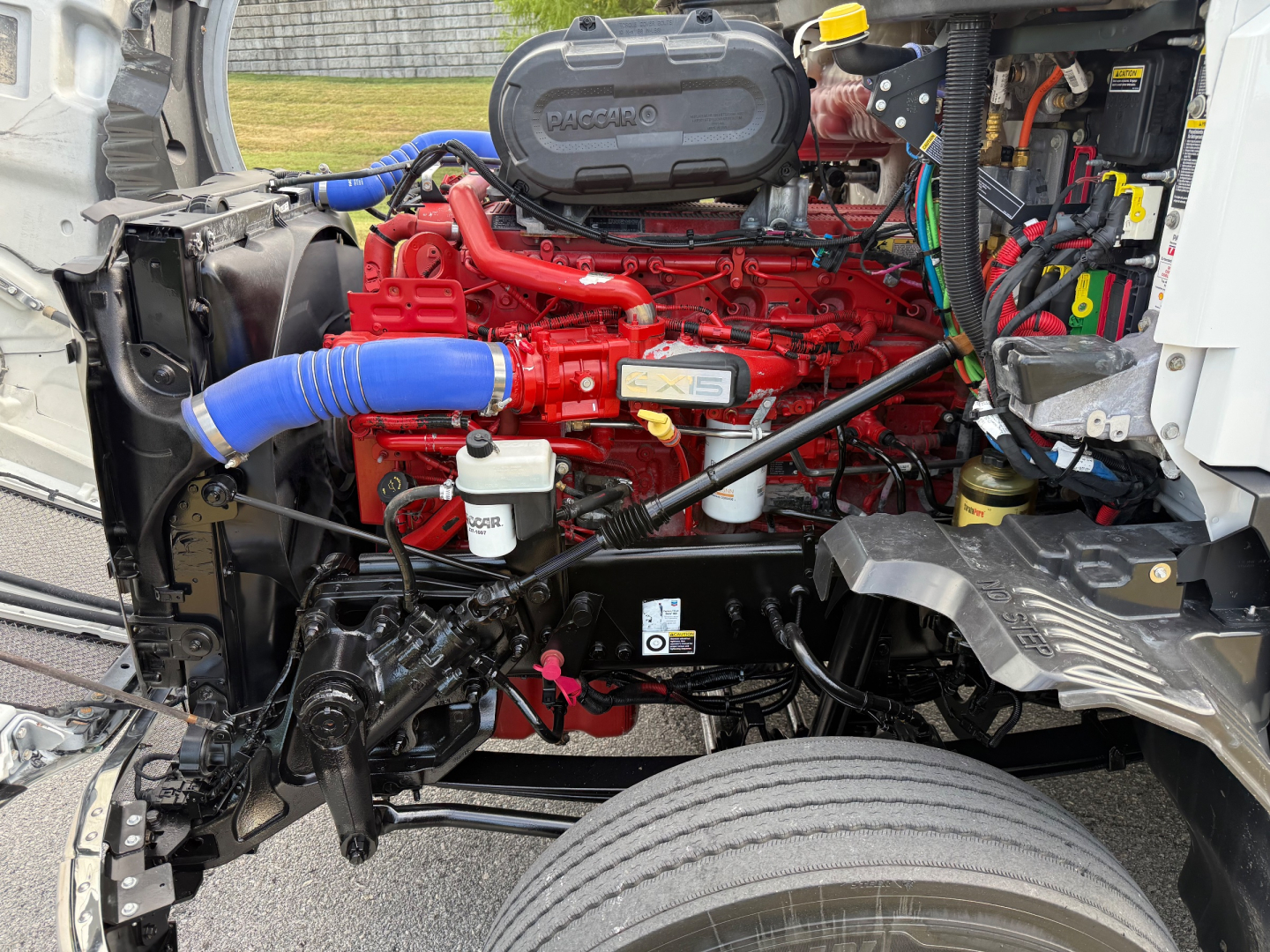 Kenworth T680 engine bay view showing red engine with blue intercooler hose, various black hoses, metal framework, and wiring harnesses.