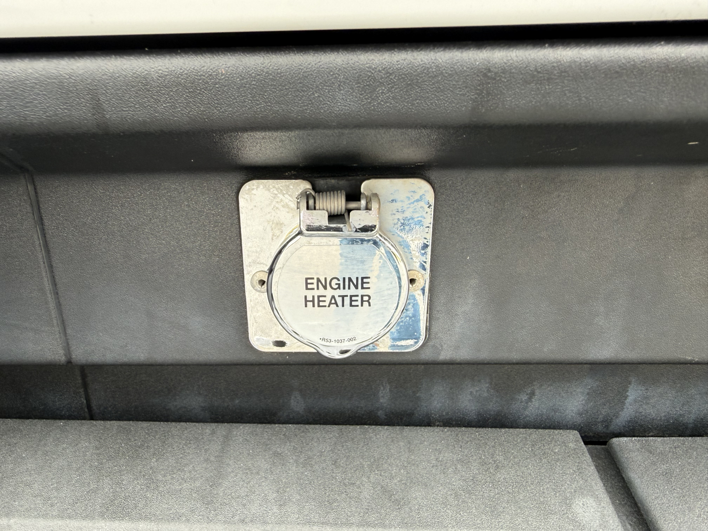 Kenworth T680 engine bay area showing a square plug-in labeled ENGINE HEATER with circular cap and surrounding metal plate.