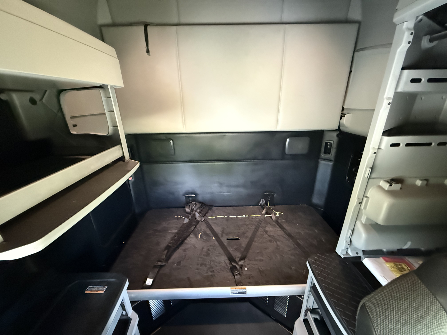 Kenworth T680 sleeper cab interior cargo area looking toward rear cab with bed and tie-downs, shelves on left, storage on right.