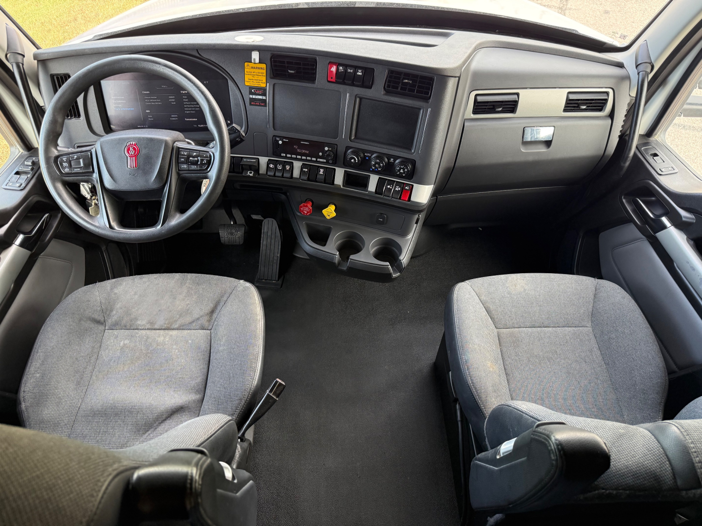 Kenworth T680 interior cab view showing steering wheel, dashboard with multiple gauges, center console, and gray fabric seats.