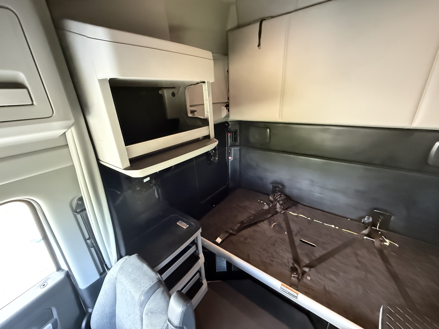 Kenworth T680 sleeper cab interior showing a mounted microwave, storage cabinet, and a folded bunk bed area with seat belt fasteners.