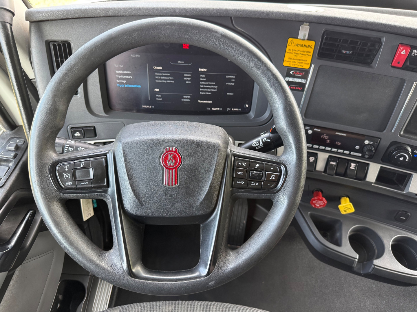Kenworth T680 cockpit showing steering wheel with Kenworth logo, digital instrument display, control buttons on wheel, and dashboard with warning labels.
