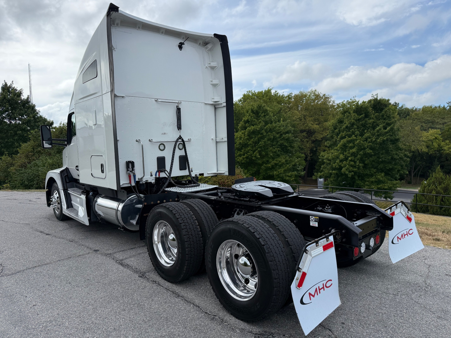 Kenworth T680 truck tractor, rear view showing dual rear axles and mud flaps with MHC logo, white sleeper cab, chrome fuel tanks, and fifth wheel area.