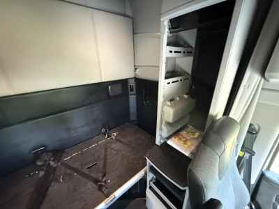 Kenworth T680 truck cab interior view showing storage compartments with shelves and a folded passenger seat; vertical cabinet with small items; light interior panels.