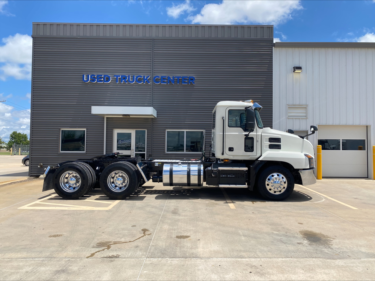 2021 Mack AN64T conventional daycab truck parked in front of Used Truck Center building. Vehicle shows three rear axles, large chrome fuel tanks, and side exhaust. Building reads 'USED TRUCK CENTER' with dark gray siding and windows.