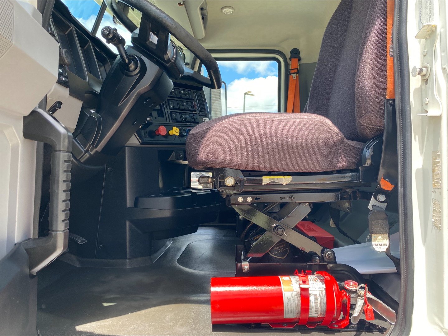 Mack AN64T truck cab interior showing driver seat, dashboard controls, steering wheel, and a red fire extinguisher mounted near floor.