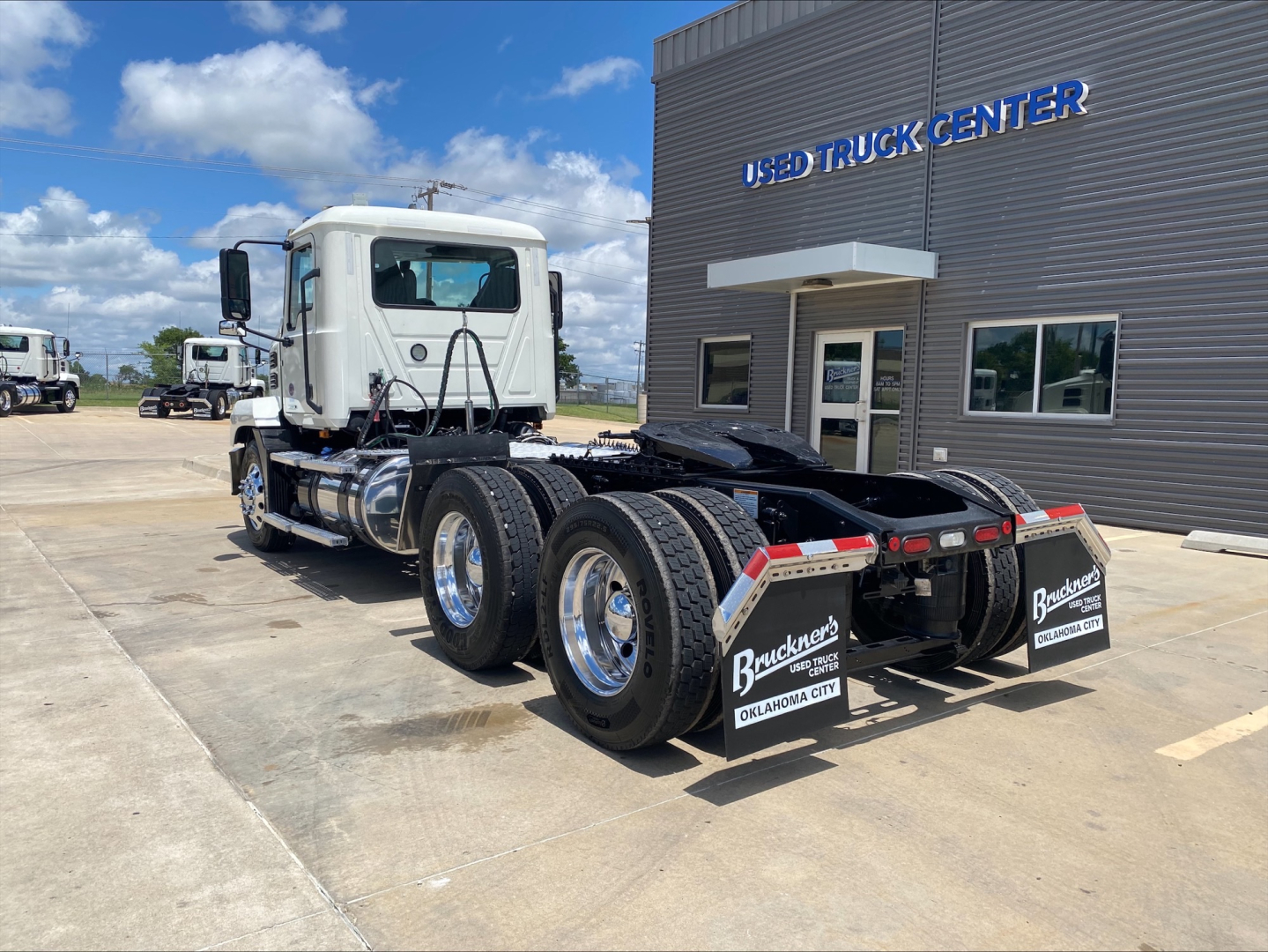 Mack AN64T conventional daycab truck in a dealership lot, rear view shows dual rear axles with mud flaps and Buckner's Oklahoma City branding. White cab, gray trailer chassis, dealership building in background.