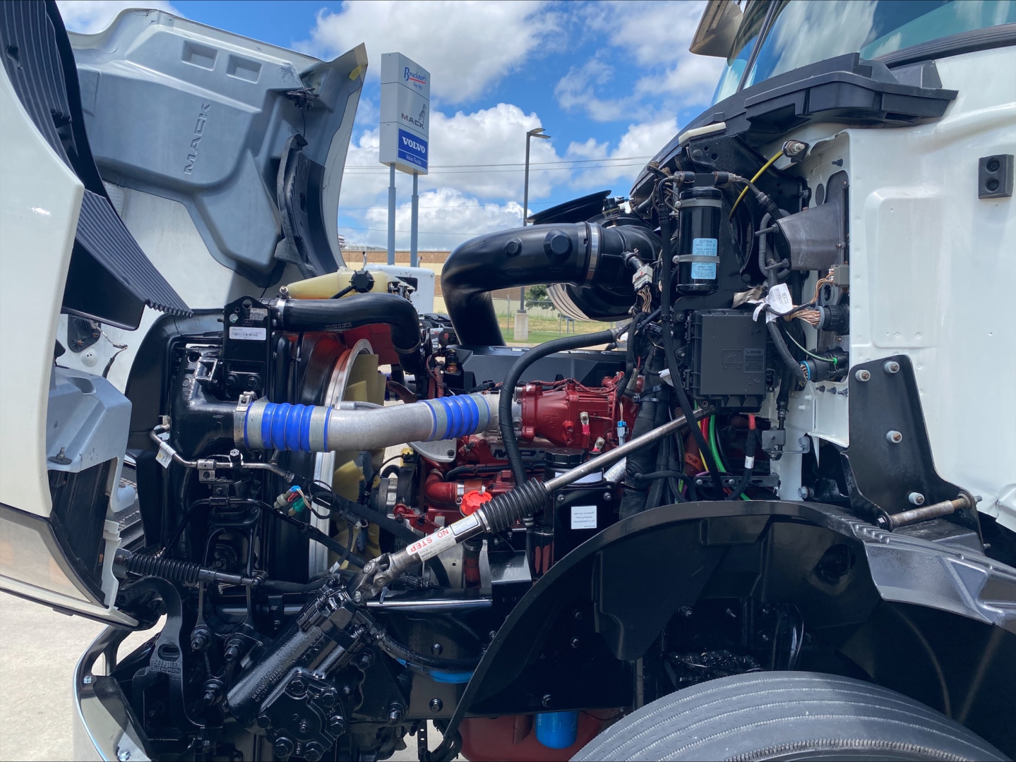 Truck engine bay of a 2021 Mack AN64T with open hood showing turbocharged diesel engine, hoses, wiring, and coolant reservoir.