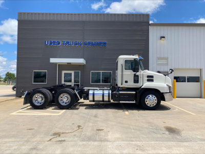 2021 Mack AN64T conventional daycab truck parked in front of Used Truck Center building. Vehicle shows three rear axles, large chrome fuel tanks, and side exhaust. Building reads 'USED TRUCK CENTER' with dark gray siding and windows.