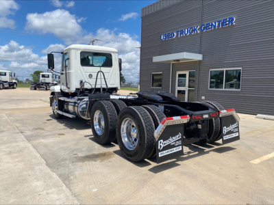 Mack AN64T conventional daycab truck in a dealership lot, rear view shows dual rear axles with mud flaps and Buckner's Oklahoma City branding. White cab, gray trailer chassis, dealership building in background.