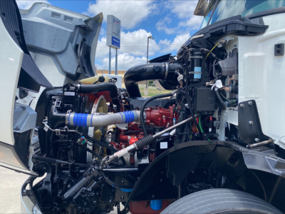 Truck engine bay of a 2021 Mack AN64T with open hood showing turbocharged diesel engine, hoses, wiring, and coolant reservoir.