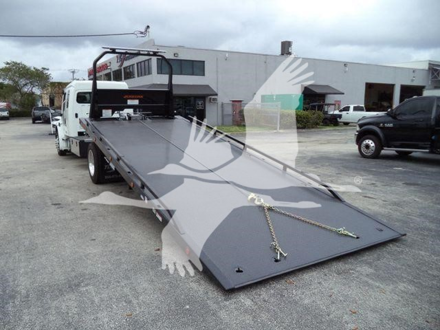 Flatbed tow truck bed extended for loading showing ramp with chains and hooks, Freightliner M2 106 in background, dealership lot.