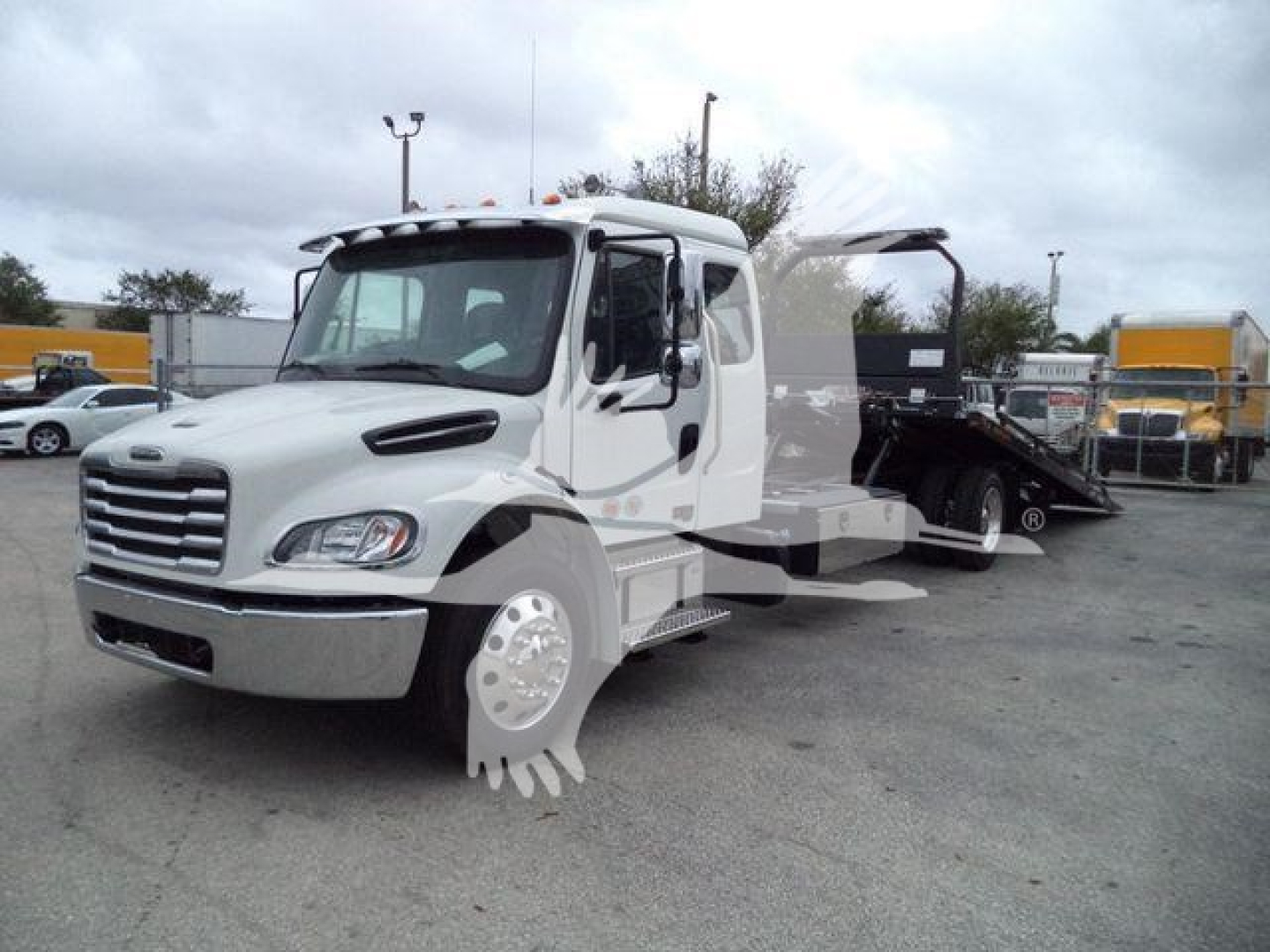Truck: Freightliner M2 106 rollback tow truck with white cab and flatbed, visible side mirrors, wheel covers, and equipment in yard.